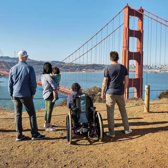 一群人, 包括一个坐轮椅的人, ist von hinten zu sehen, während sie von den Marin Headlands aus auf die Golden Gate Bridge blicken.